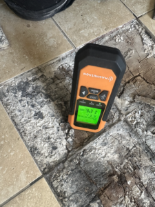 Moisture-meter reading recorded on the exposed floor/substrate following localised removal of the tiled finish. Use this image to show the investigation progressing beneath the visible tiled surface