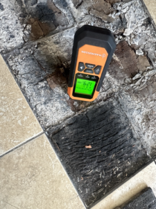 Moisture meter positioned over the exposed deteriorated timber/substrate. This providess supporting evidence that the concealed floor materials were being assessed as part of the investigation.