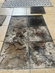 Exposed bathroom floor following removal of tiles, revealing extensive deterioration to the timber substrate. The exposed timber is severely degraded and fragmented, with areas of darkened material, loss of section and deteriorated bedding/adhesive visible across the floor area. This is the strongest representative image of the concealed defect and should be the principal investigation photograph